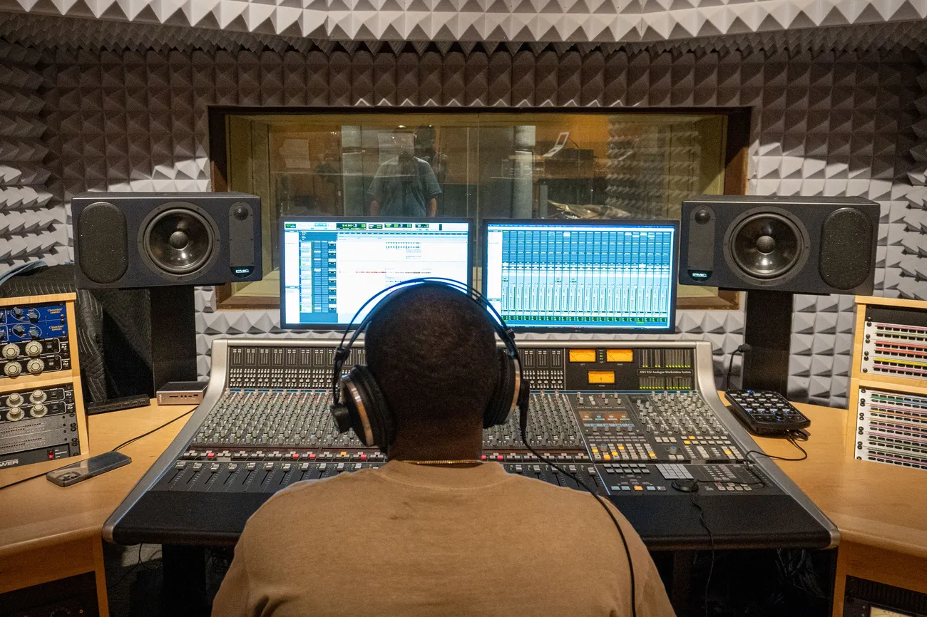 A person sits at a console in a recording studio. The person's back is to the camera.