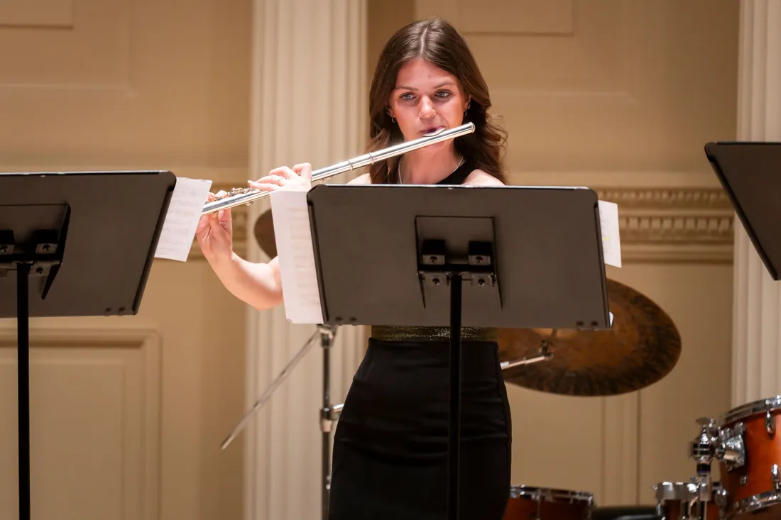 A person stands behind a music stand and plays a flute formally dressed.
