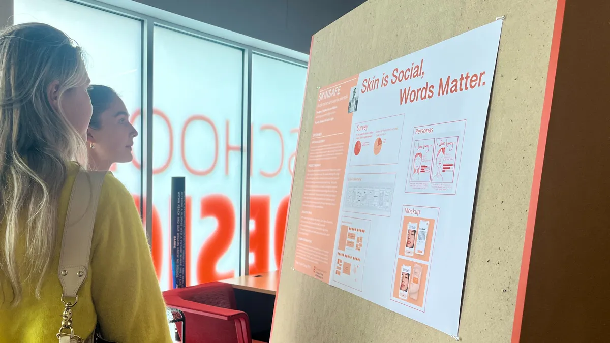 Two students look at an easel that is displaying a poster that reads Skin is Social, Words Matter.