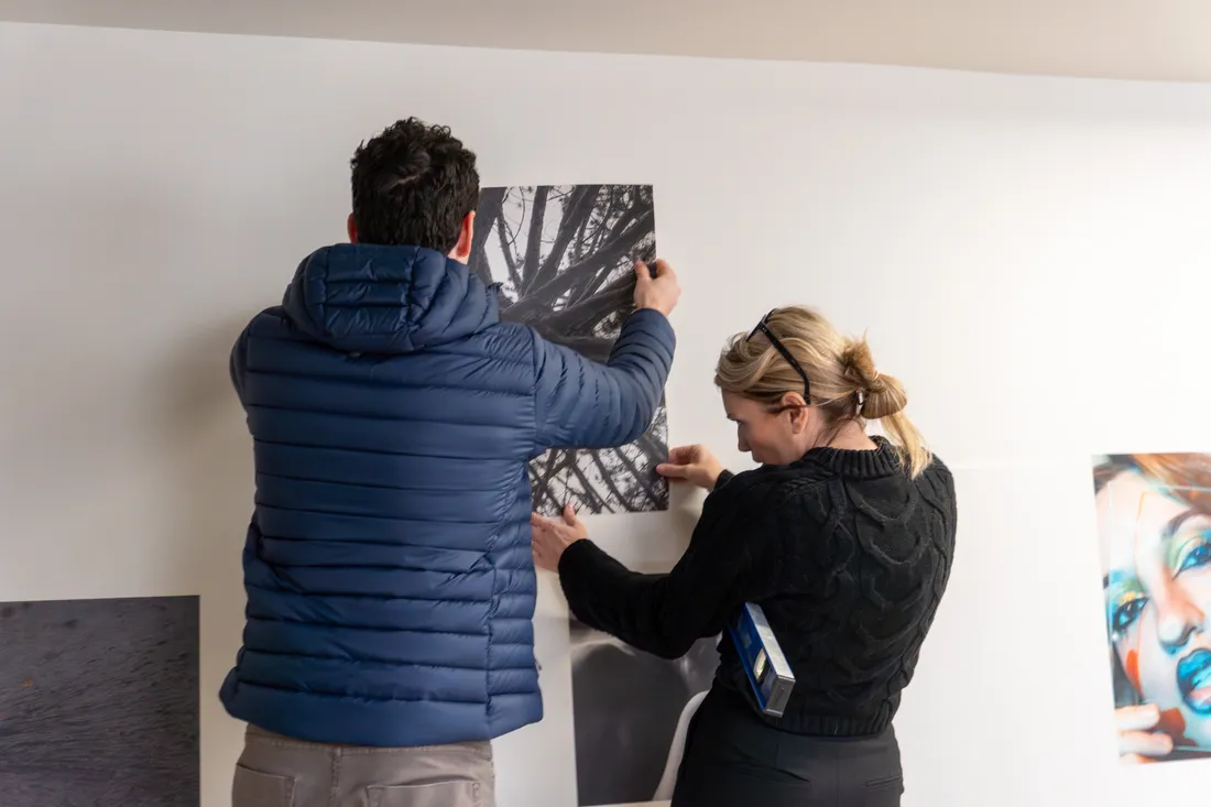 Two people hang photographs on a wall.