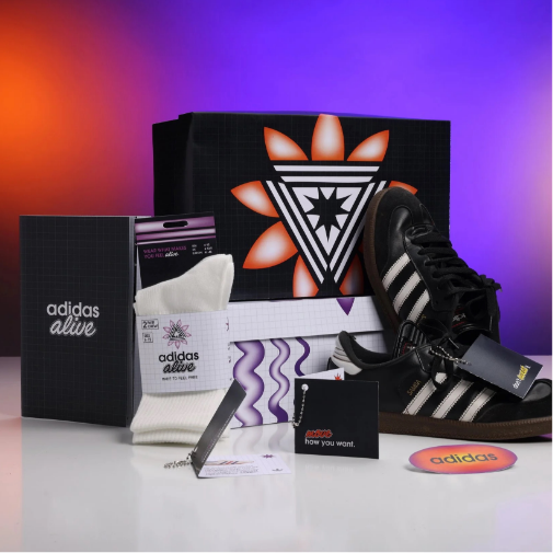 A branded Adidas promotional gift set displayed against a vibrant purple-to-orange gradient background, featuring black Adidas sneakers, socks, packaging boxes, printed cards, and accessories with bold modern graphics and “adidas alive” branding.