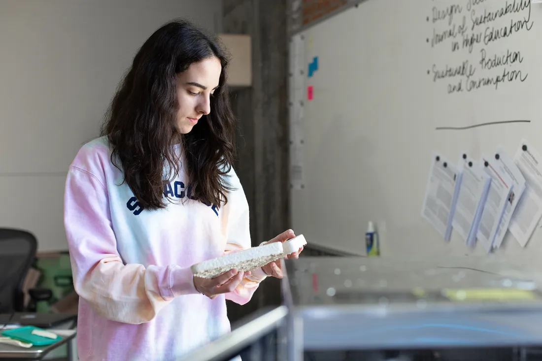 A student holds a small piece of a white material in her hands standing next to a white board with writing on it.
