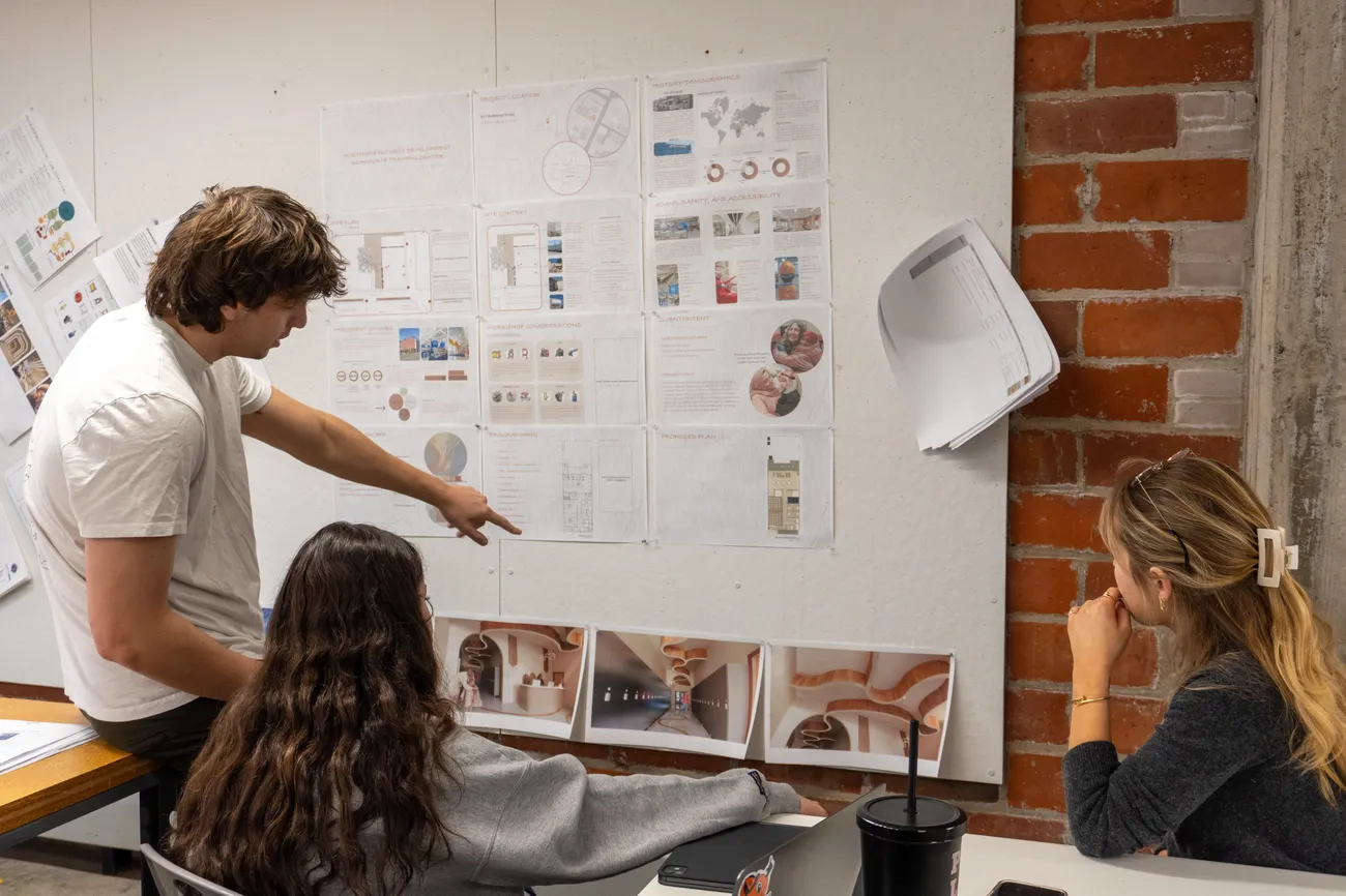 Three students sit in front of a white wall with pictures of designs pinned to it. They are discussing the designs.