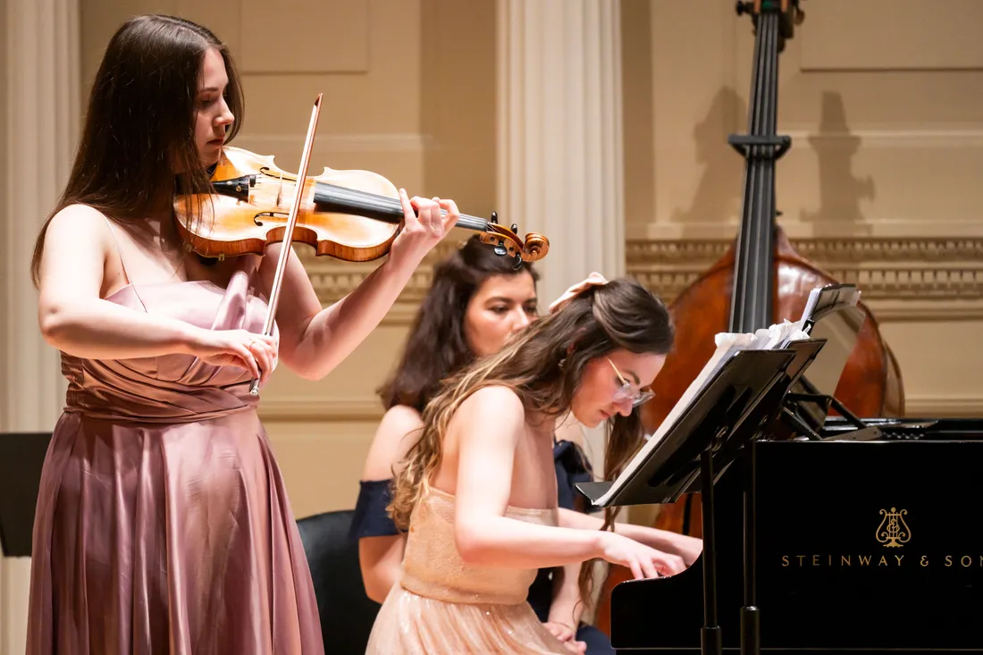 A student stands and plays a violin next to a student seated at and playing a piano. Both are dressed in formal attire.