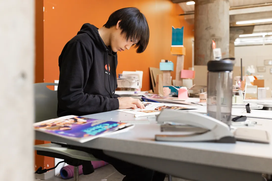 A student stands at a table working on assembling a booklet. On the table is a water bottle and pages of the booklet.