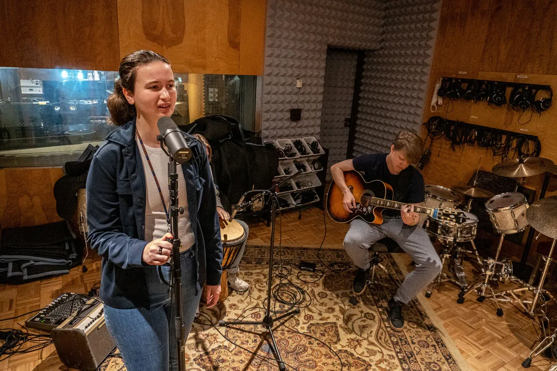 A person stands at a microphone in a recording studio. A person sits and plays the guitar in the background.