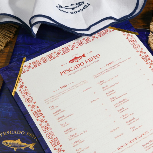 A neatly designed seafood restaurant menu for “Pescado Frito” displayed on a wooden table, featuring red decorative borders and sections for fish, chips, and house-made sauces. Surrounding the menu are branded blue-and-white napkins and packaging with a nautical theme.