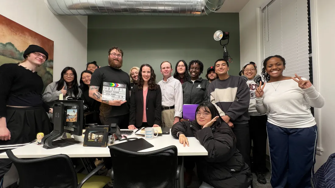 A group of individuals pose in a room. Jack Auger holds a clapperboard in the center.