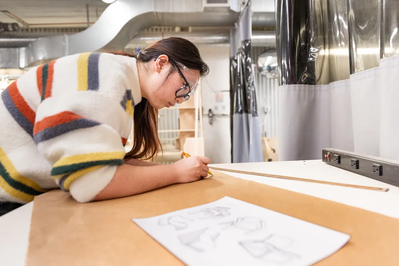 A student leans over a table and draws with a pencil on brown paper. A drawing on white paper sits on top of the brown paper.