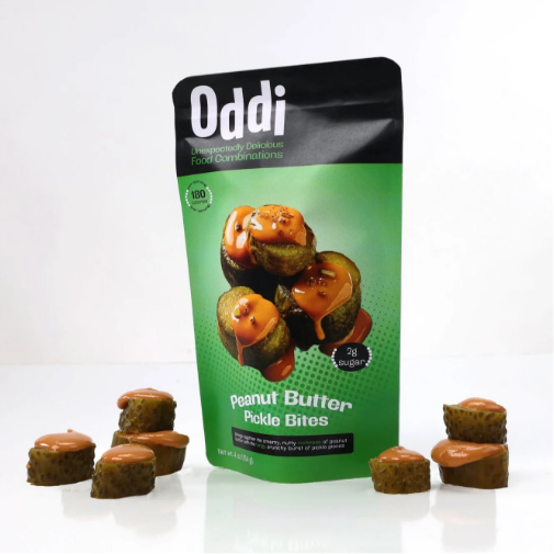 A bold green snack package labeled “Oddi Peanut Butter Pickle Bites” stands against a white background, featuring images of pickle slices topped with peanut butter drizzle. Several pickle bite snacks are arranged around the package, highlighting the unusual sweet-and-savory flavor combination.