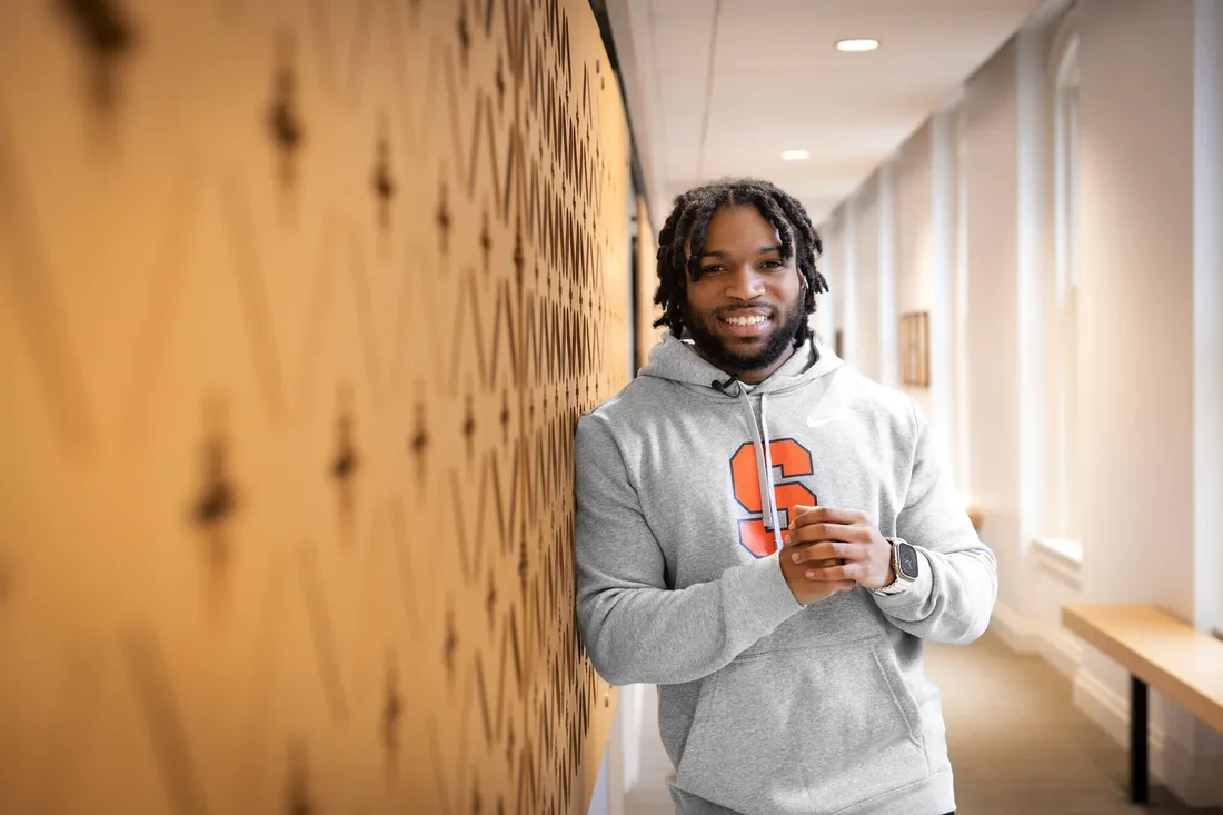 A student leans agains a wall in a hallway and smiles.