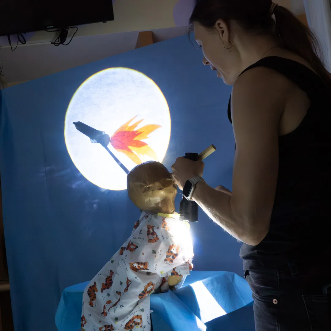 A puppet stares at a screen that shows a rocket crossing a moon.