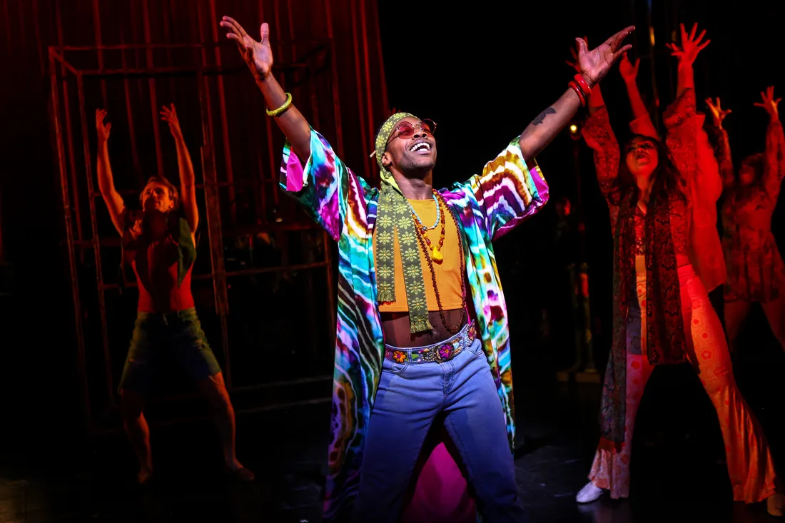 An actor in jeans and a tie-dye shirt raises his hands and sings.
