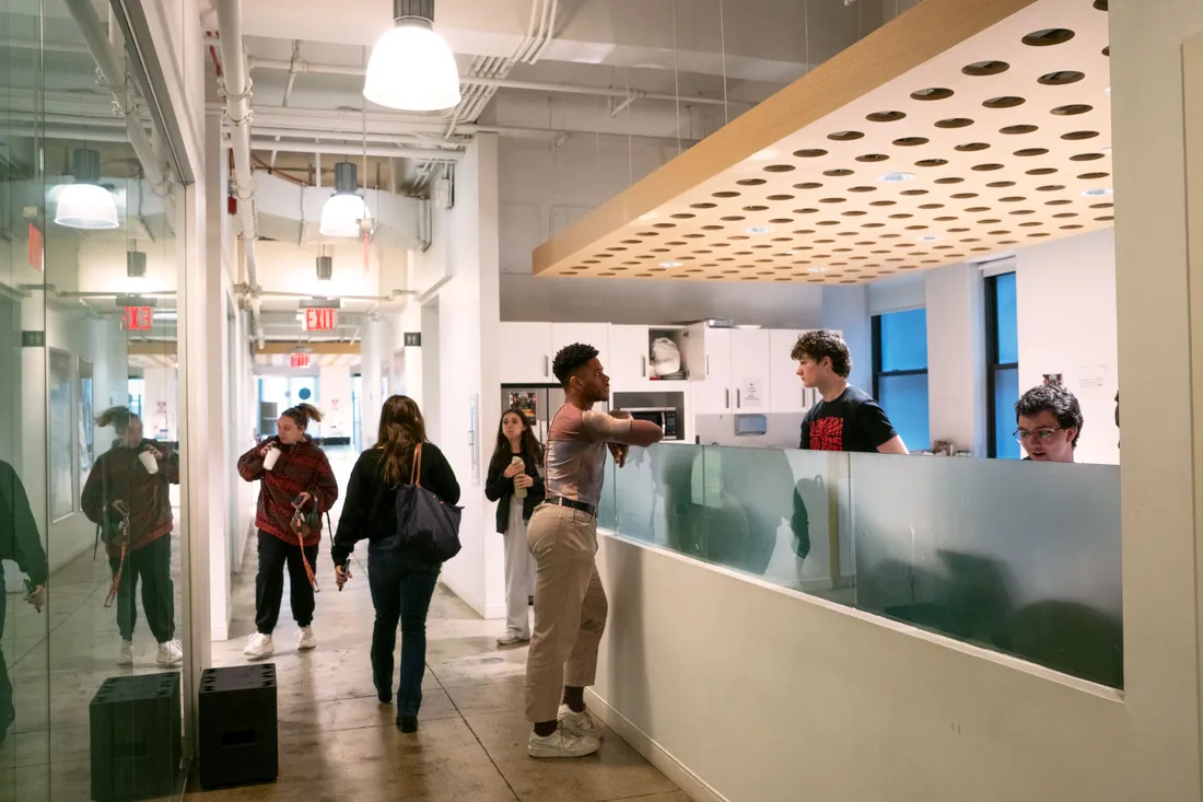 People walk down a hallway. On one side of the hallway is a reception desk where people are working and talking.