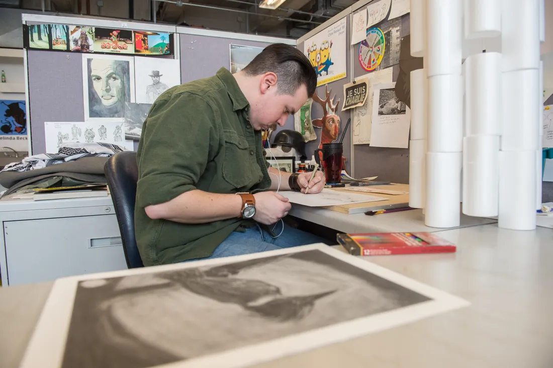 A student sits at a table in a studio drawing on a piece of paper.