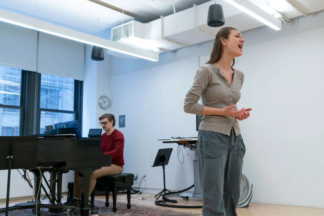 A student sings in a rehearsal studio. In the background a person plays piano.