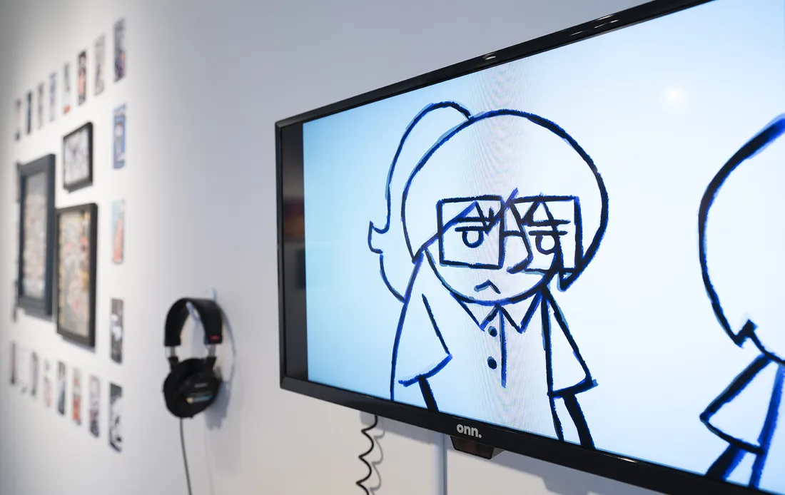 A video screen on a gallery wall shows a cartoon-like drawing of a character wearing a short-sleeved shirt and glasses.