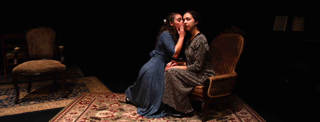 Two actors in costume sit on a dimly lit stage. One whispers to the other.