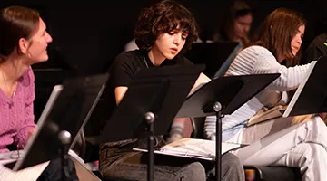 Three students sit at music stands with black binders and scripts. One of the students makes a note on the script with a pen.
