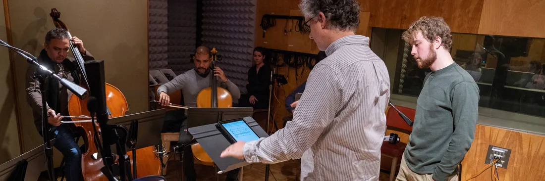 A teacher and student stand at a music stand in a recording studio at a music stand while two string players play in the background.