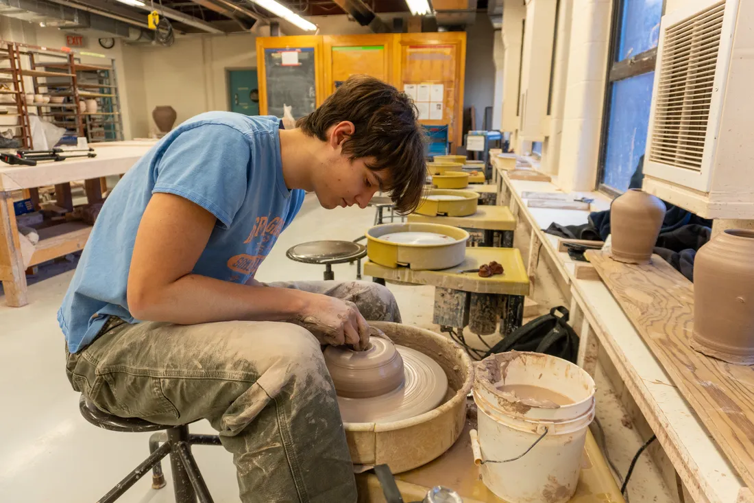A student sits at a pottery wheel spinning a lump of clay.