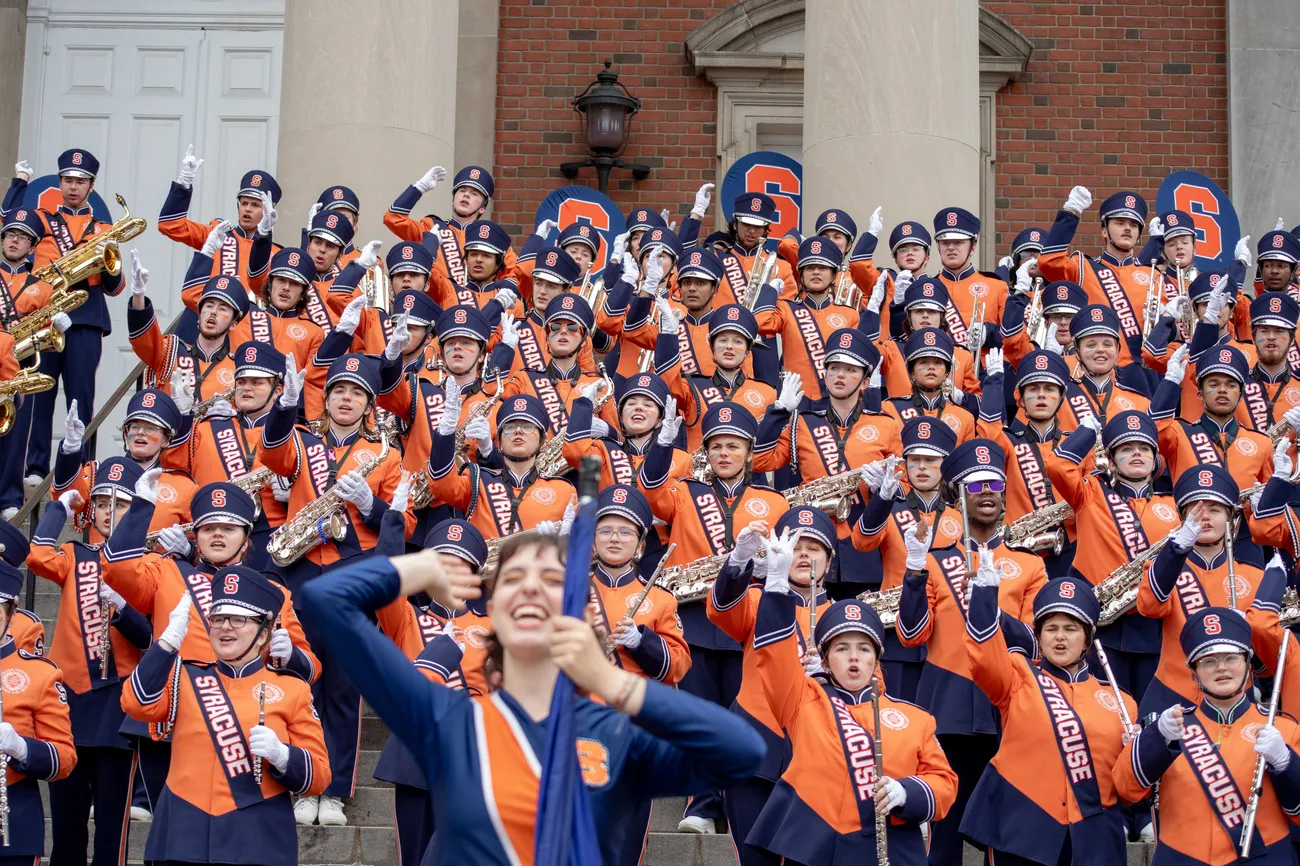 Members of a marching band perform in a group dressed in orange and blue uniforms.