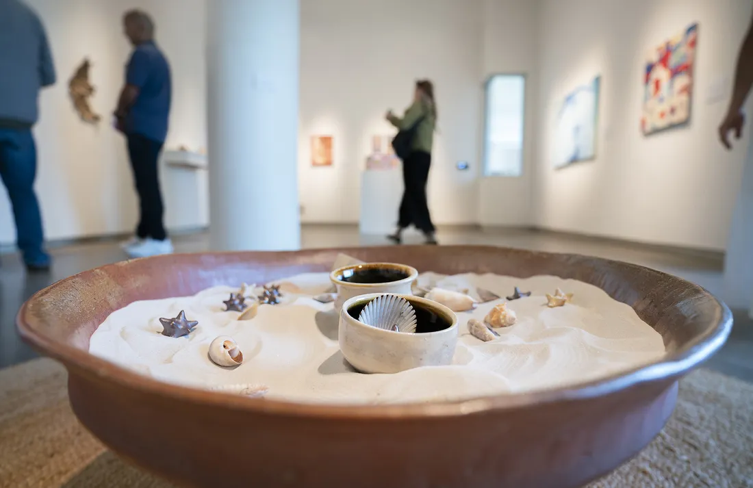 A ceramic bowl of sand and shells sits on the floor of an art gallery. In the background, people look at artwork on the walls of the gallery.