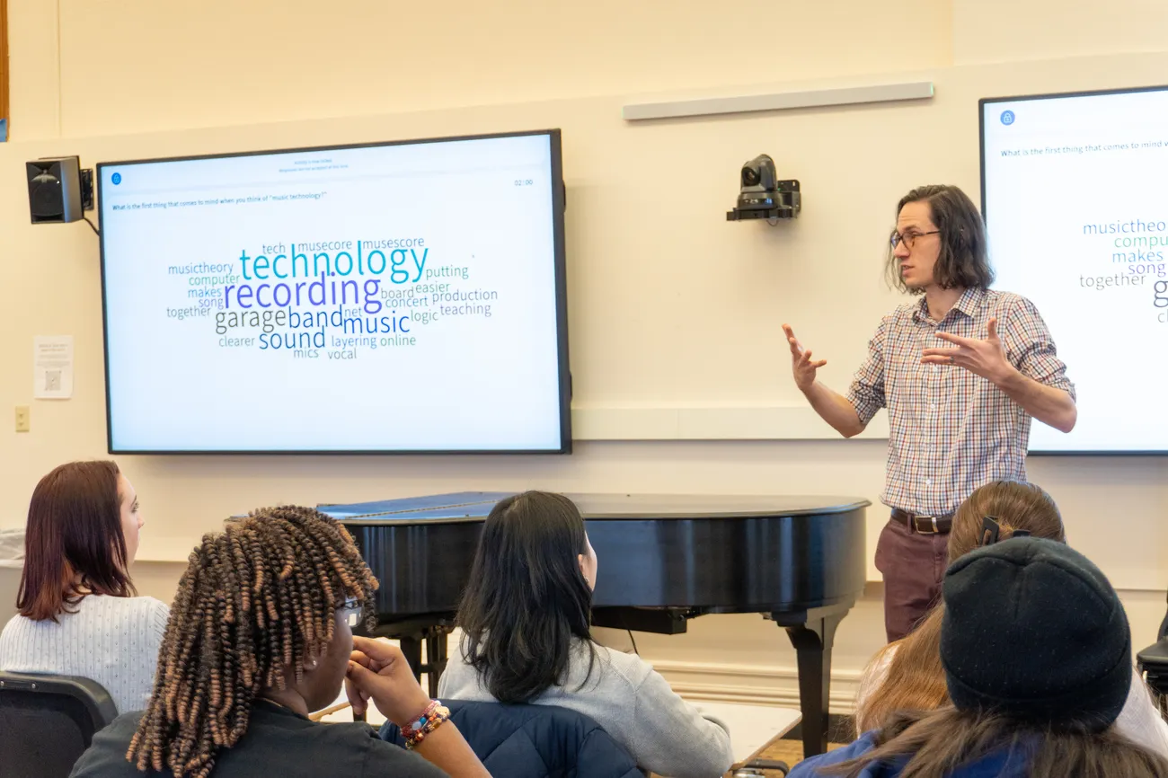 A faculty member stands at the front of a classroom gesturing next to a screen on the wall that contains class information.