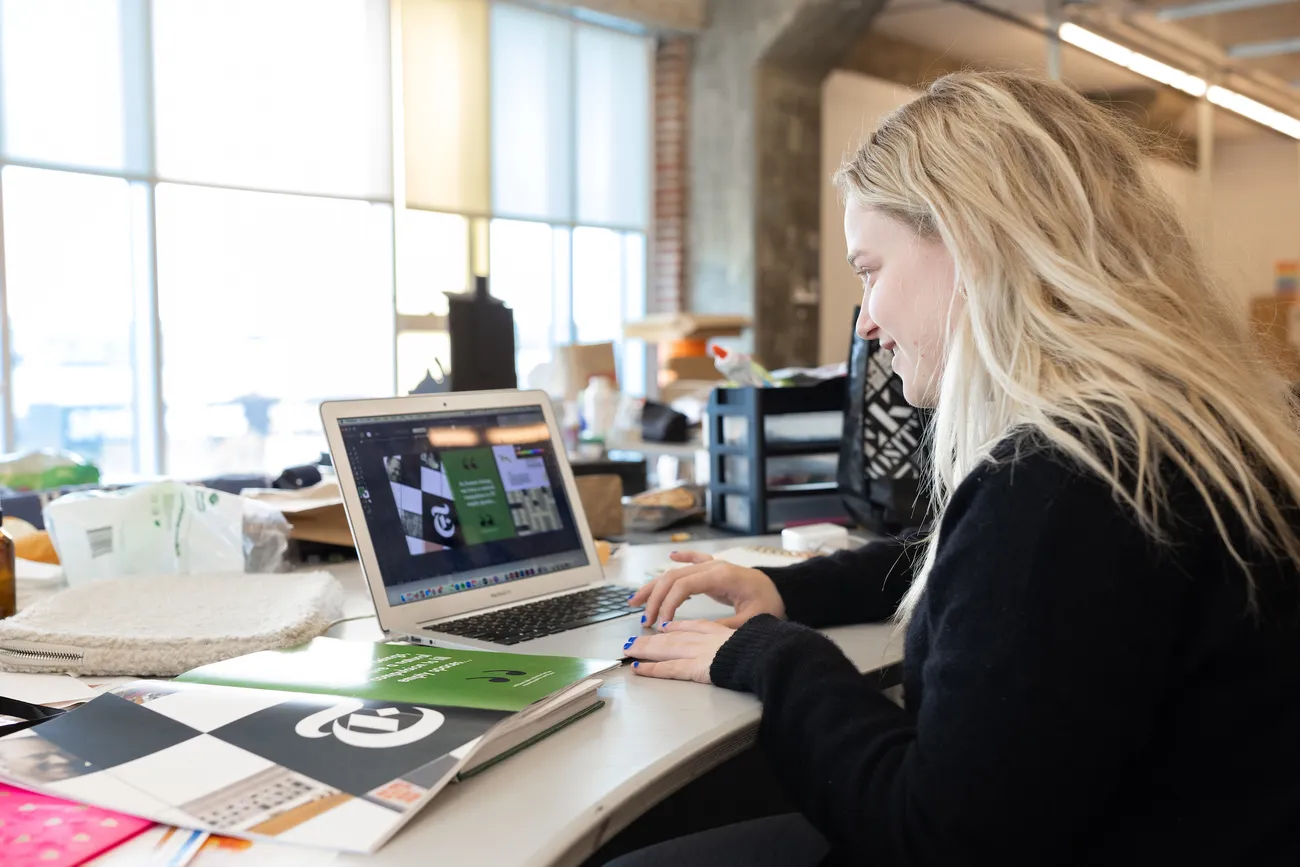A student sits at a desk looking at a laptop. Next to the student are printed papers with graphic design on them.