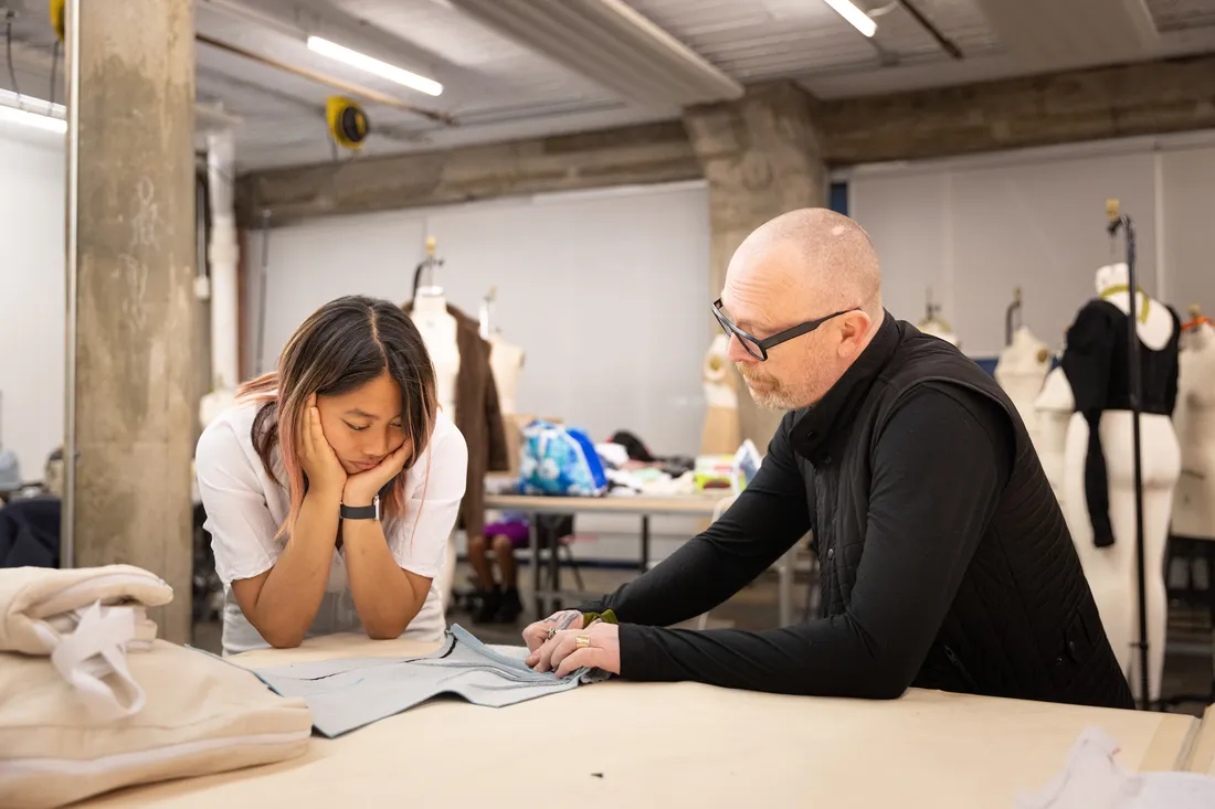 A faculty member sits at a table with a student and looks at a piece of fabric on the table.
