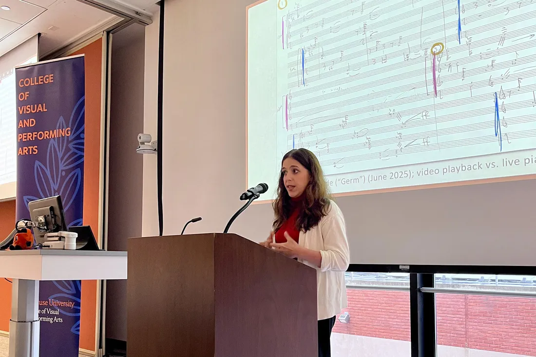 Loren Loiacono stands at a lectern speaking into a microphone. Behind her is a large screen displaying a music score.