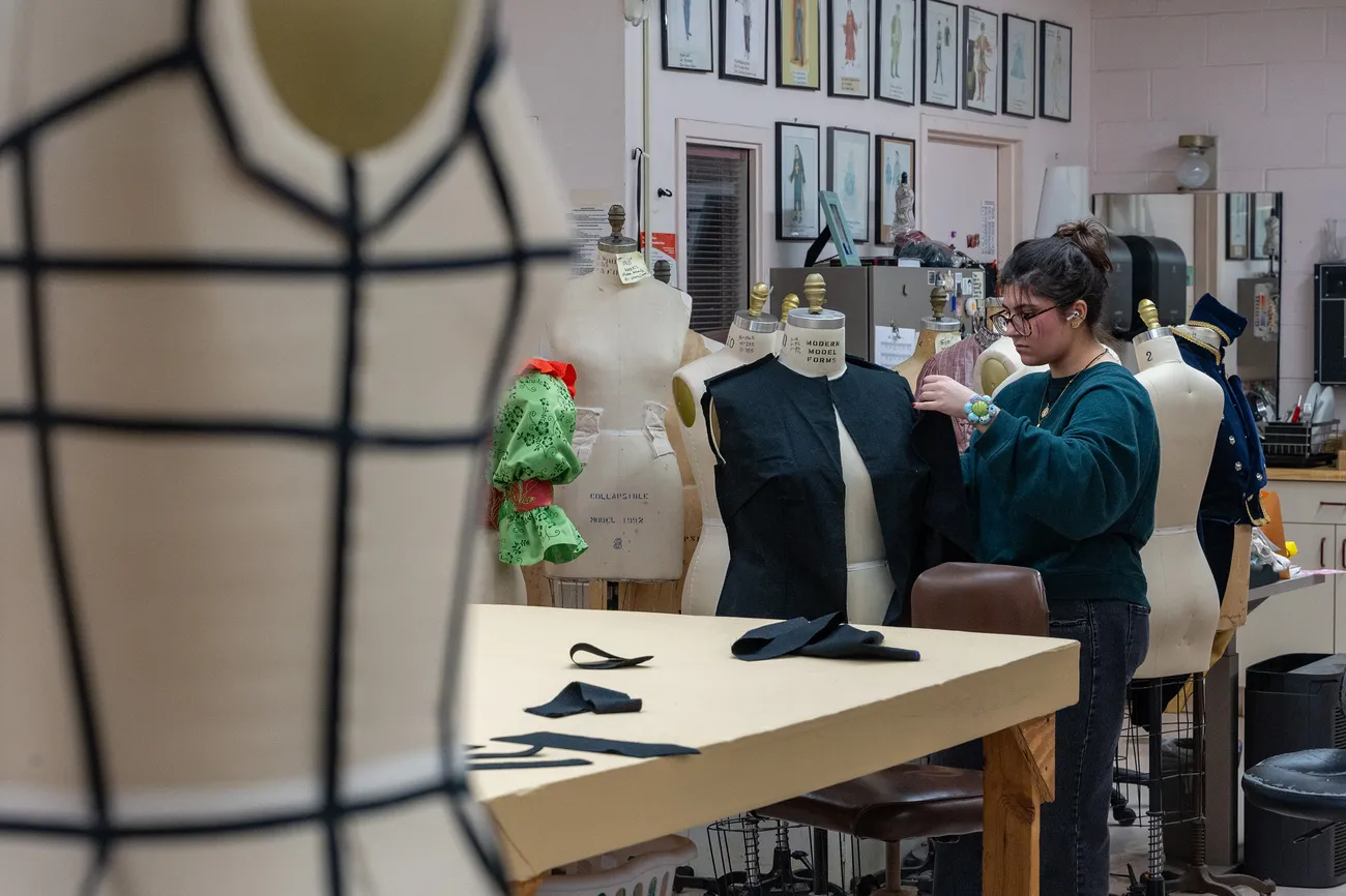 A student sews a costume on a mannequin in a studio surrounded by mannequins and drawings of costumes.