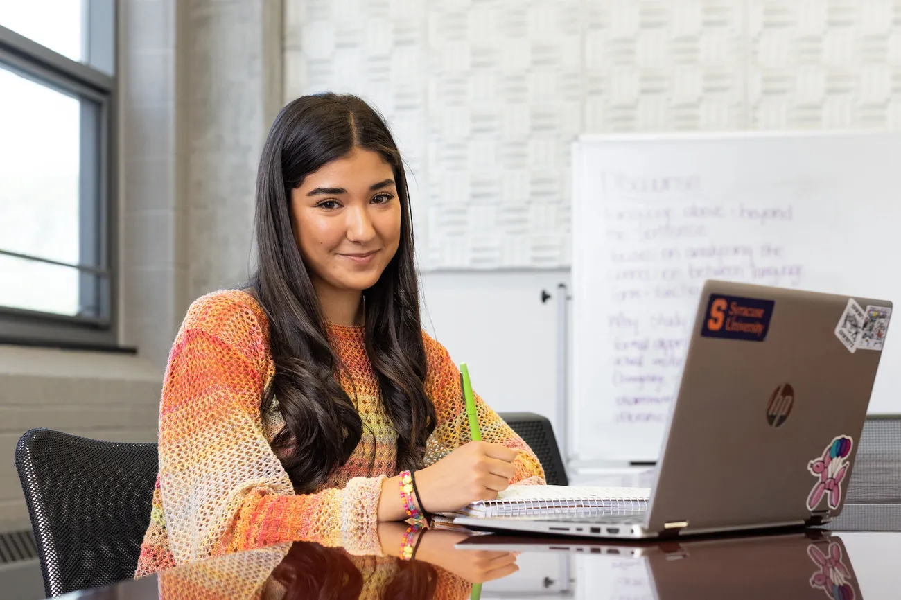 A student sits at a table in a classroom working on a laptop.