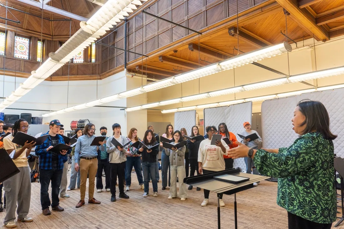 A choir rehearses in a room led by a conductor.