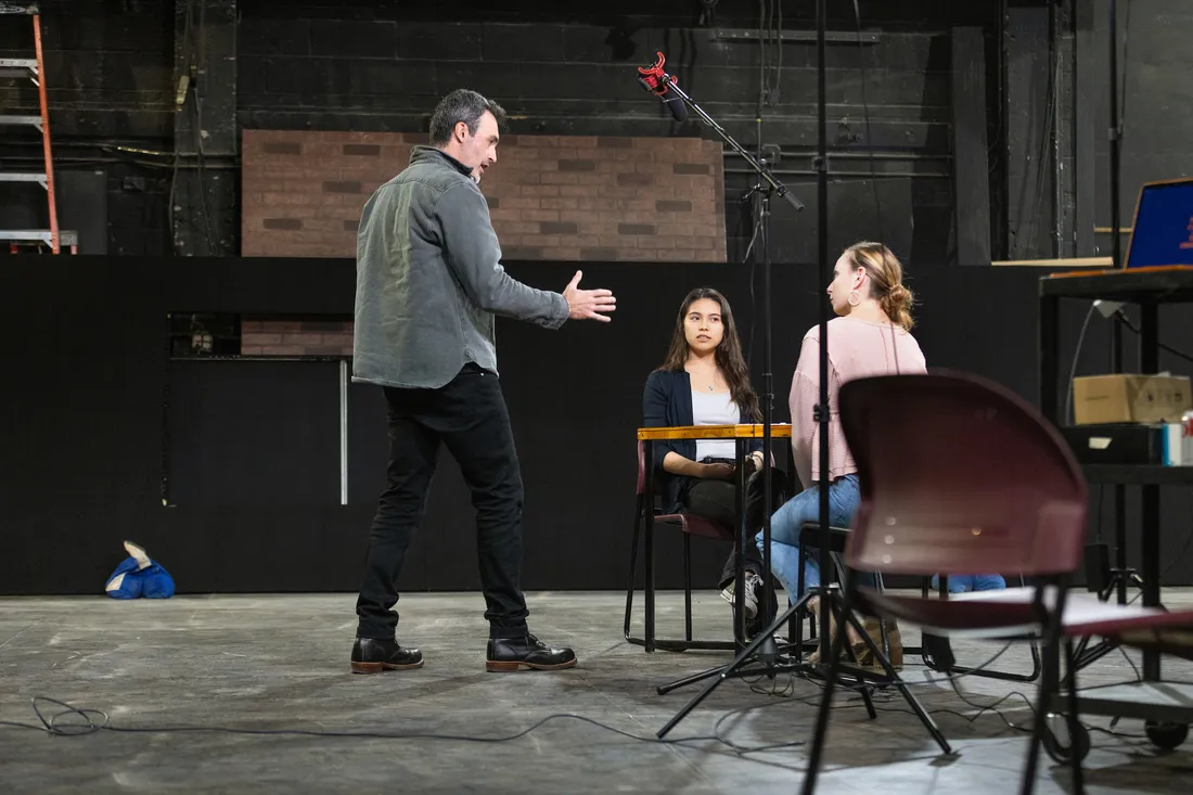 A person stands on a stage and talks to two people who are seated at a table on the stage.