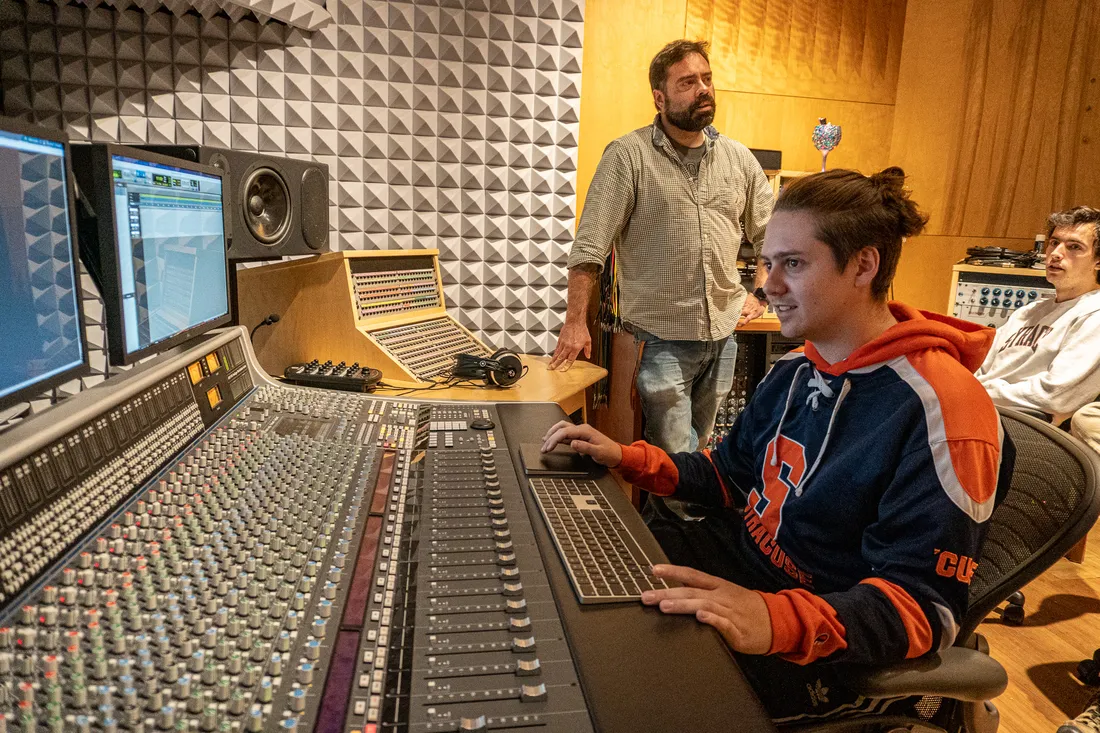 A student sits at a recording console in a recording studio. A person stands behind the student in the background.