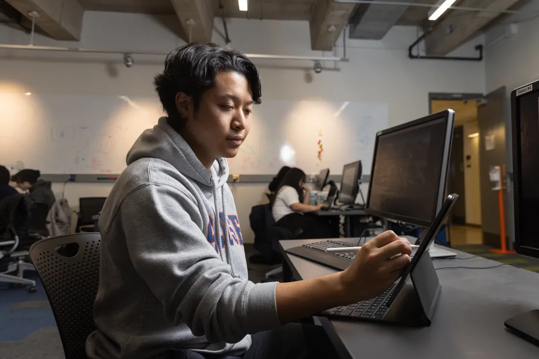 A student sits in front of a computer screen and uses a tablet next to the screen.