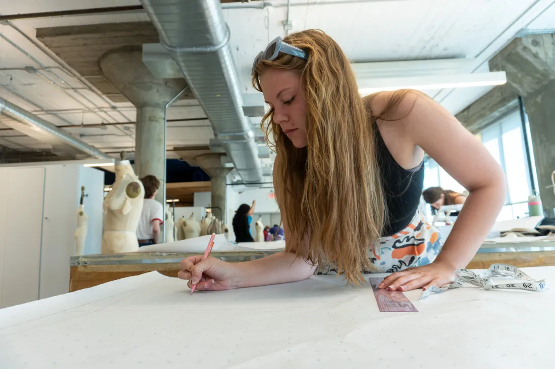 A student leans over a table drawing. There is a ruler and measuring tape on the table.