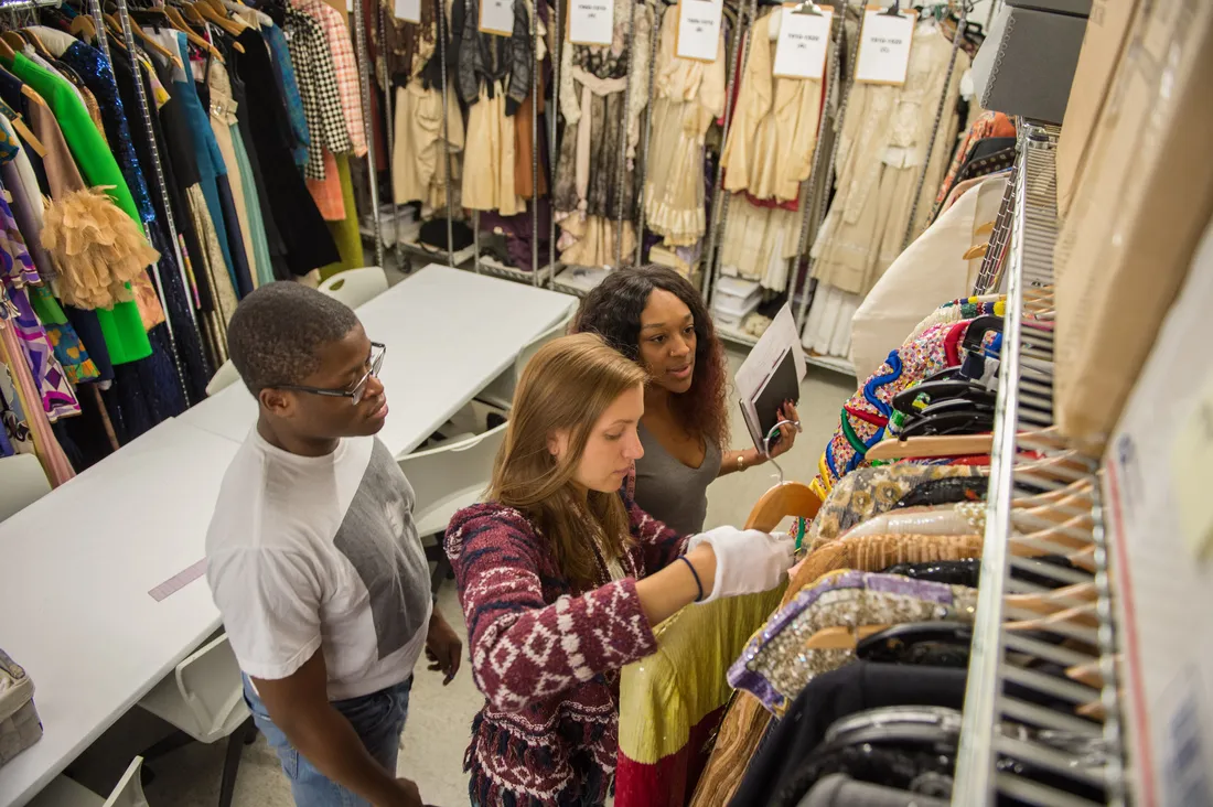 Three students stand among racks of clothing. One student has pulled an item of clothing from a rack.