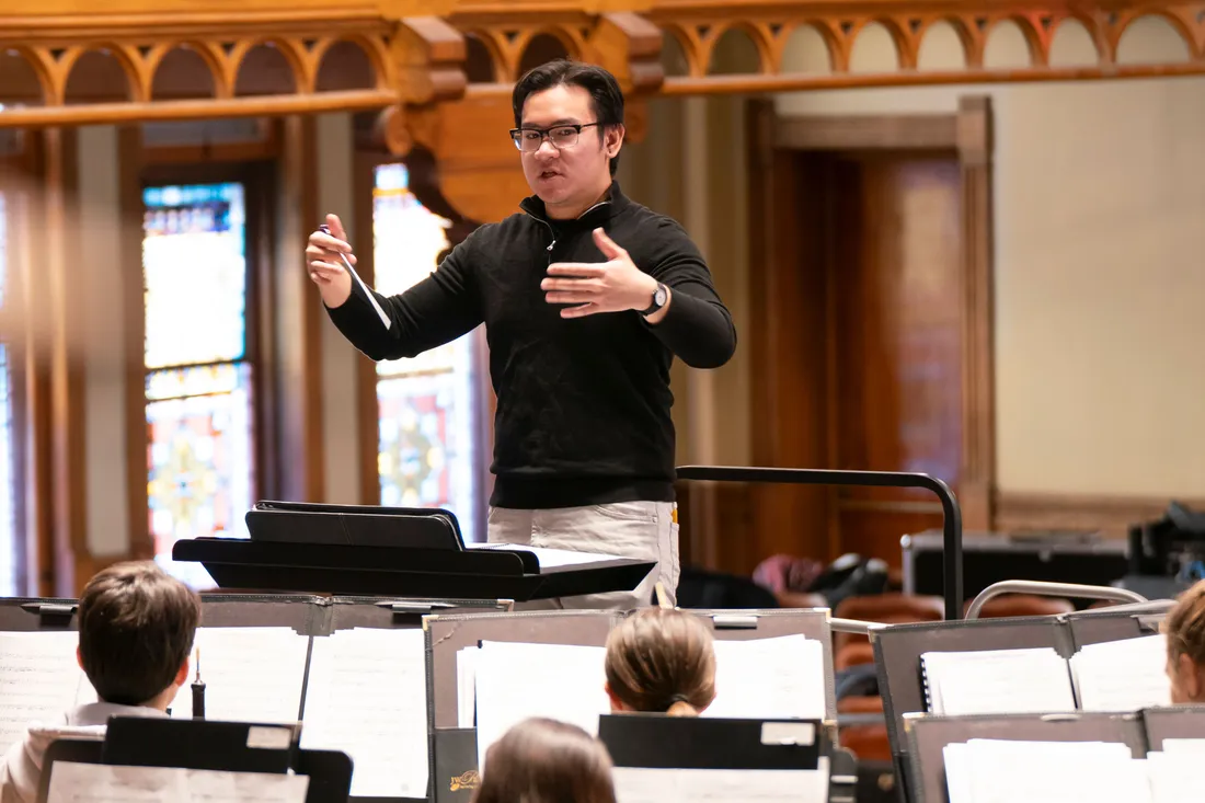 A student conductor stands in front of student musicians and conducts them in a rehearsal.