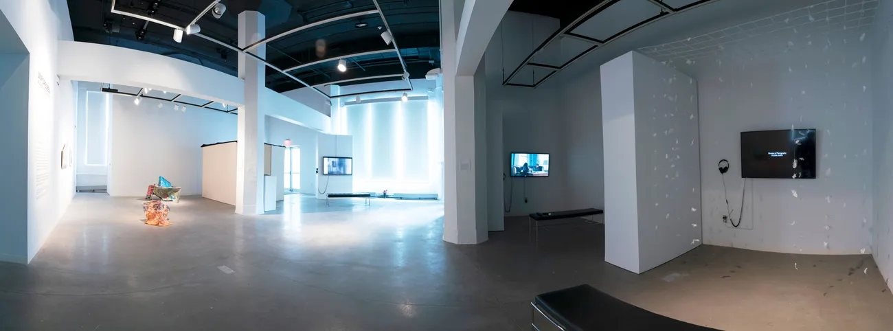 A wide shot of a gallery divided into different sections with artwork on screens and sculpture in the middle of the floor.