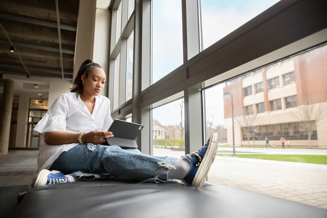 A student sits on a bench next to a window looking at a tablet.