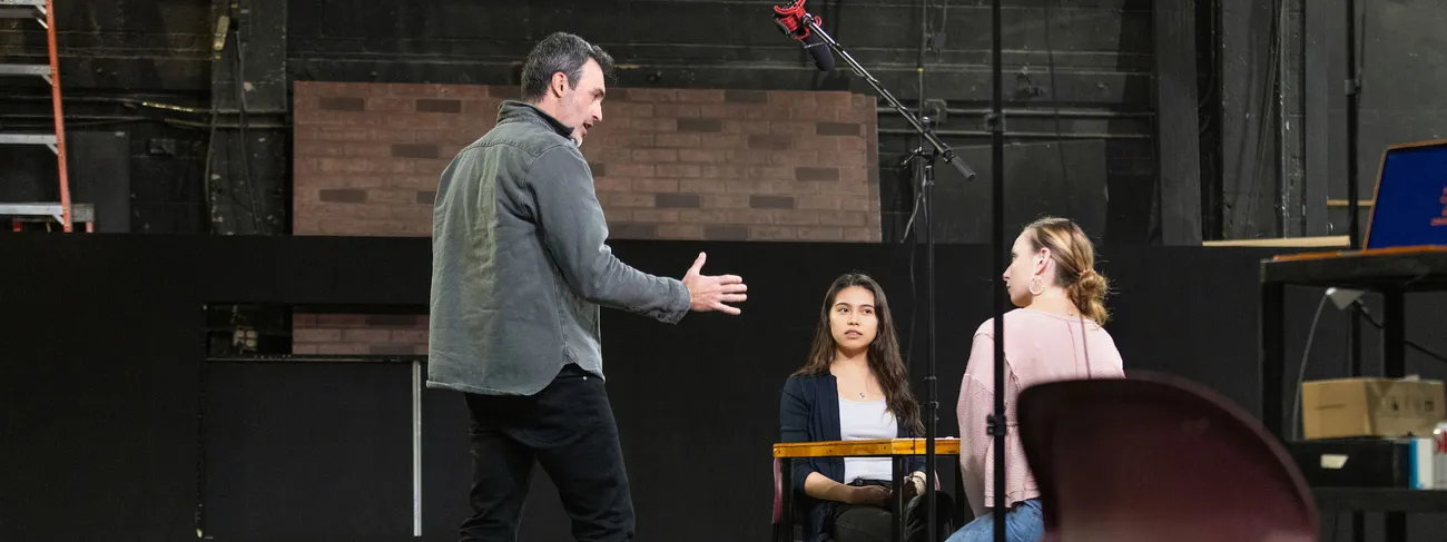 A man talks to two students seated on a stage.
