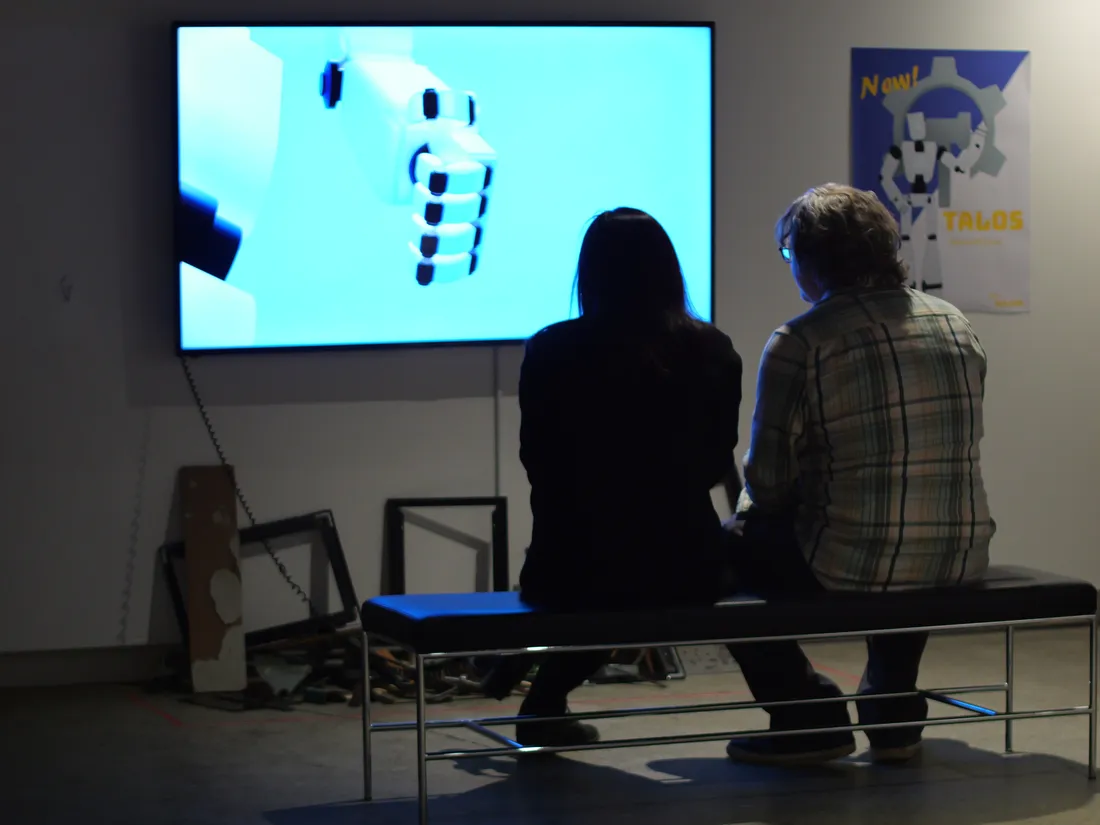 Two people sit on a bench in a gallery looking at a screen with computer-generated images.