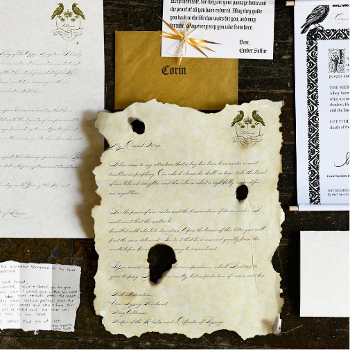 A collection of vintage-style stationery and handwritten documents arranged on a dark wooden surface, featuring aged parchment with burnt edges and wax seals, decorative envelopes, elegant calligraphy, and ornate black-and-white printed invitations with a historical aesthetic.