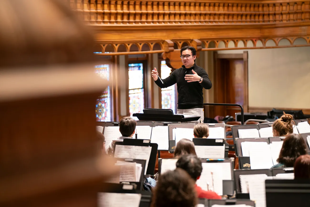 A person stands in front of a wind ensemble holding a conducting baton with hands raised.