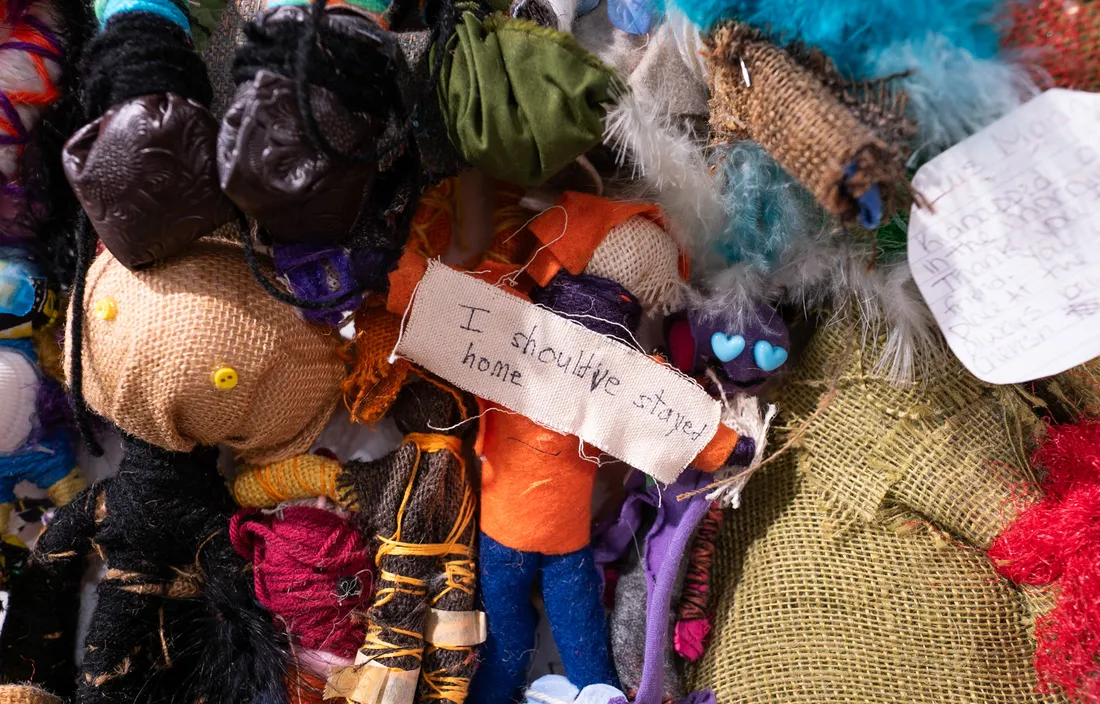 An assortment of handmade dolls. A strip of fabric in the center reads, “I should’ve stayed home.”