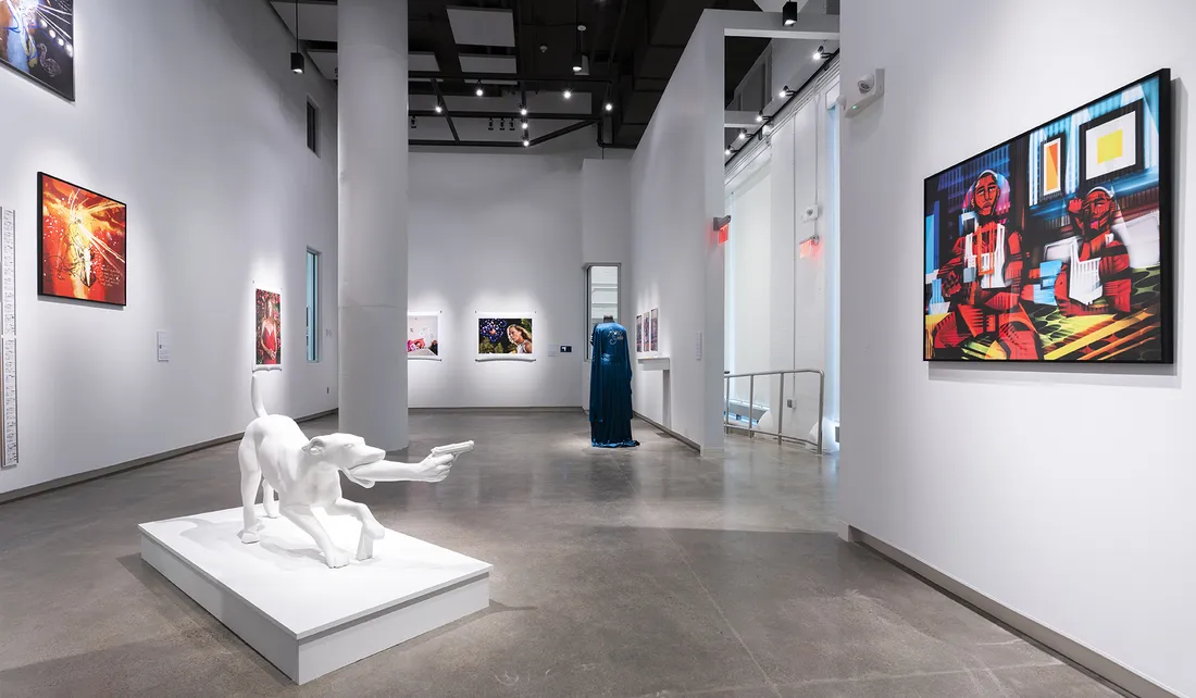 A gallery with paintings and photographs on the wall. In the middle of the gallery floor is a sculpture and a mannequin wearing a robe.