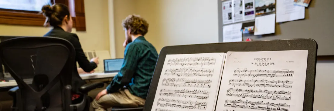 A music stand with music on it. In the background are two people sitting at a desk looking at a laptop.