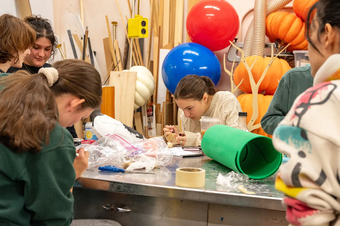 A group of students sit and work at a table in a workshop surrounded by theater props.